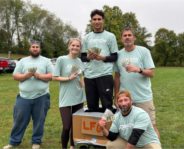Five JDB NOW team members in matching seafoam shirts celebrating outdoors after a team competition